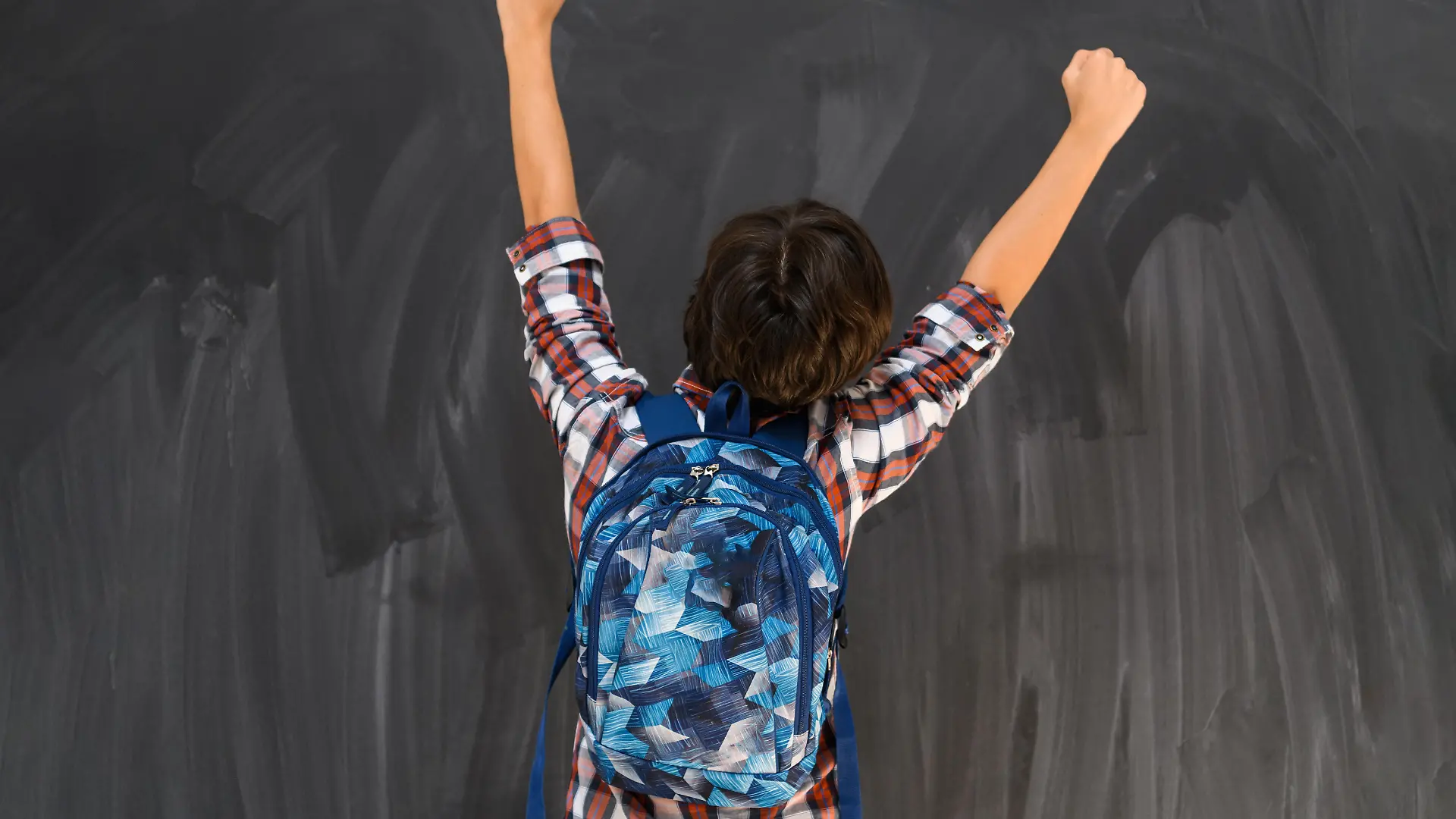 Junge mit dunklen Haaren und Schulrucksack steht vor einer Tafel mit dem Rücken zum Betrachter und reißt die Arme hoch.