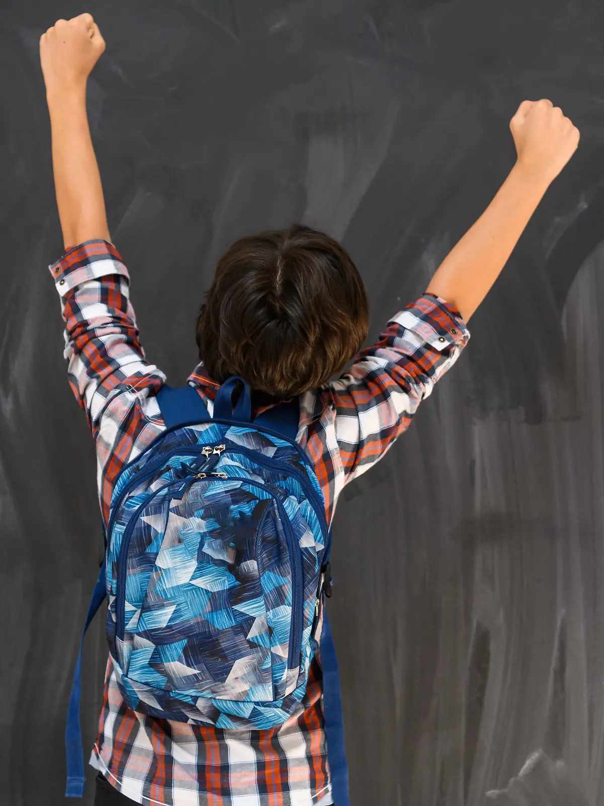 Junge mit dunklen Haaren und Schulrucksack steht vor einer Tafel mit dem Rücken zum Betrachter und reißt die Arme hoch.