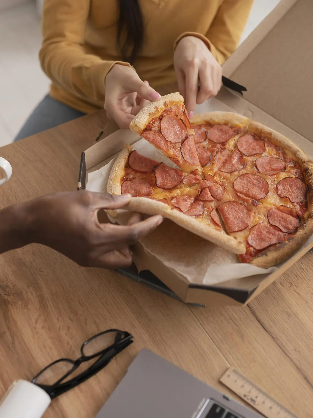 Zwei Hände greifen auf einem Tisch nach Salamipizza in einem Karton
