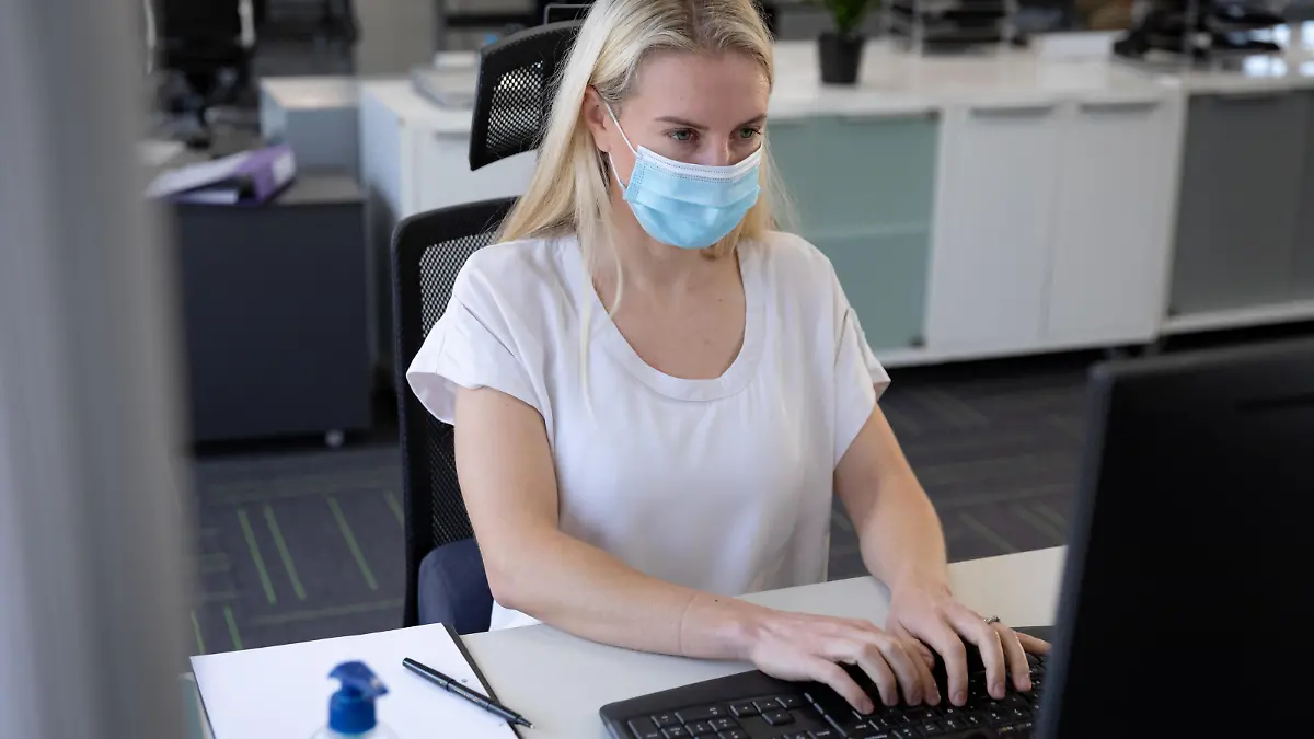 Caucasian businesswoman working in a casual office wearing face mask, sitting at desk using computer. Hygiene and social distancing in workplace during Coronavirus Covid 19 pandemic. || Modellfreigabe vorhanden