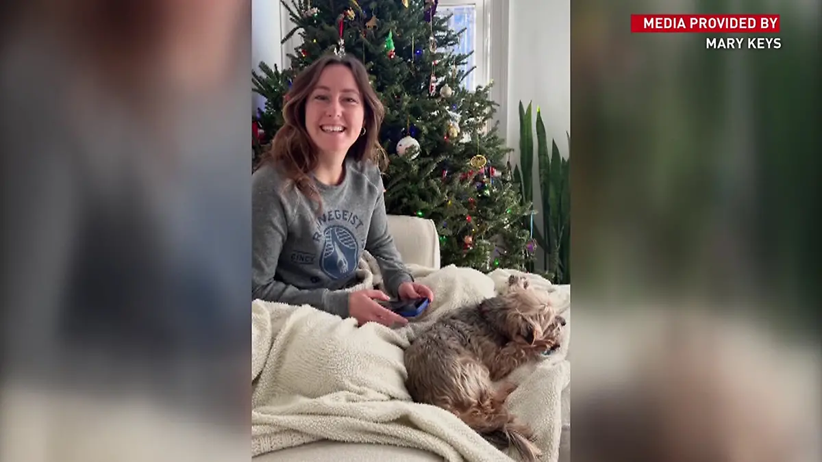 Eine junge Frau liegt mit einem Hund auf dem Sofa und blickt in die Kamera.