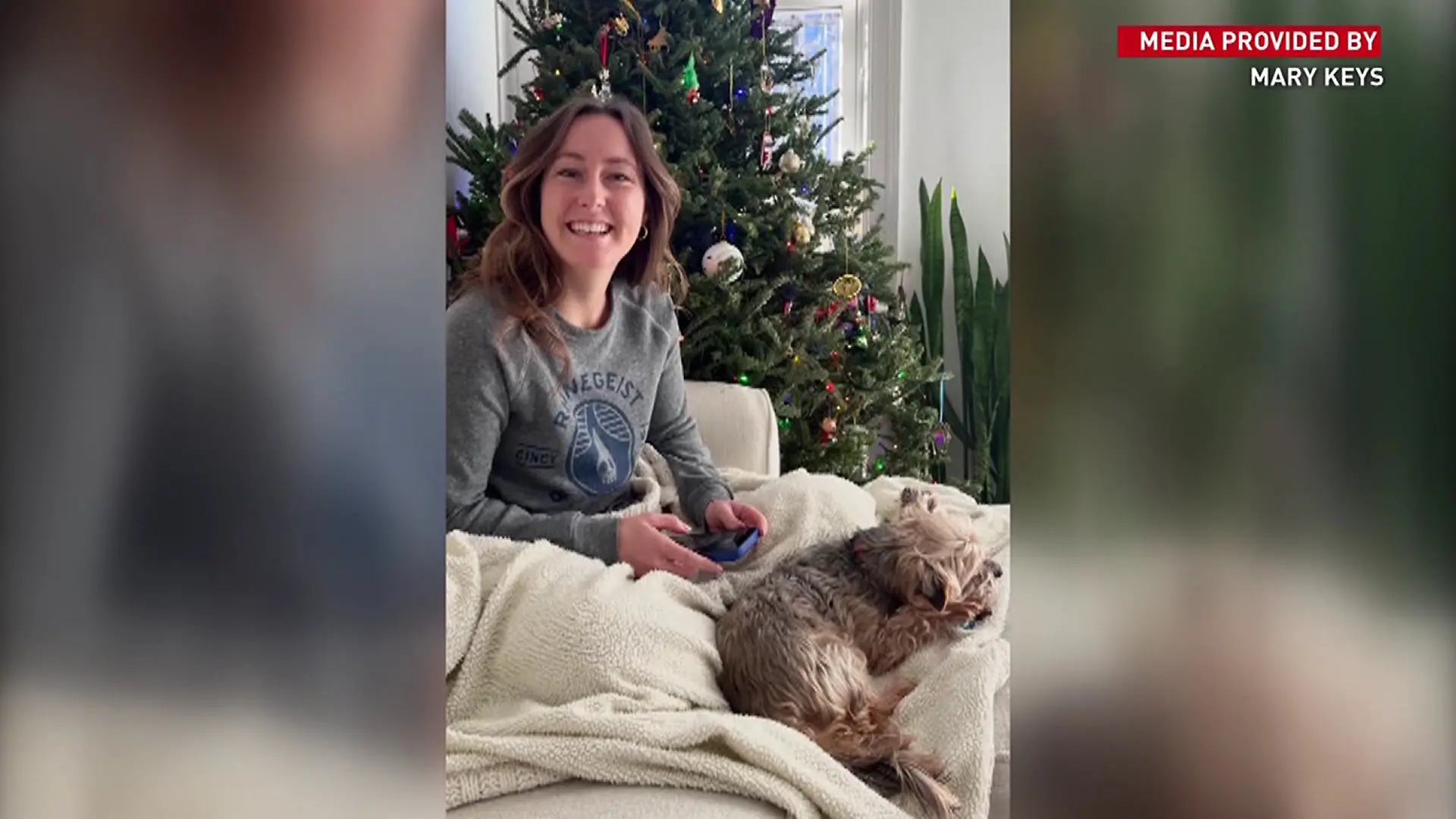 Eine junge Frau liegt mit einem Hund auf dem Sofa und blickt in die Kamera.