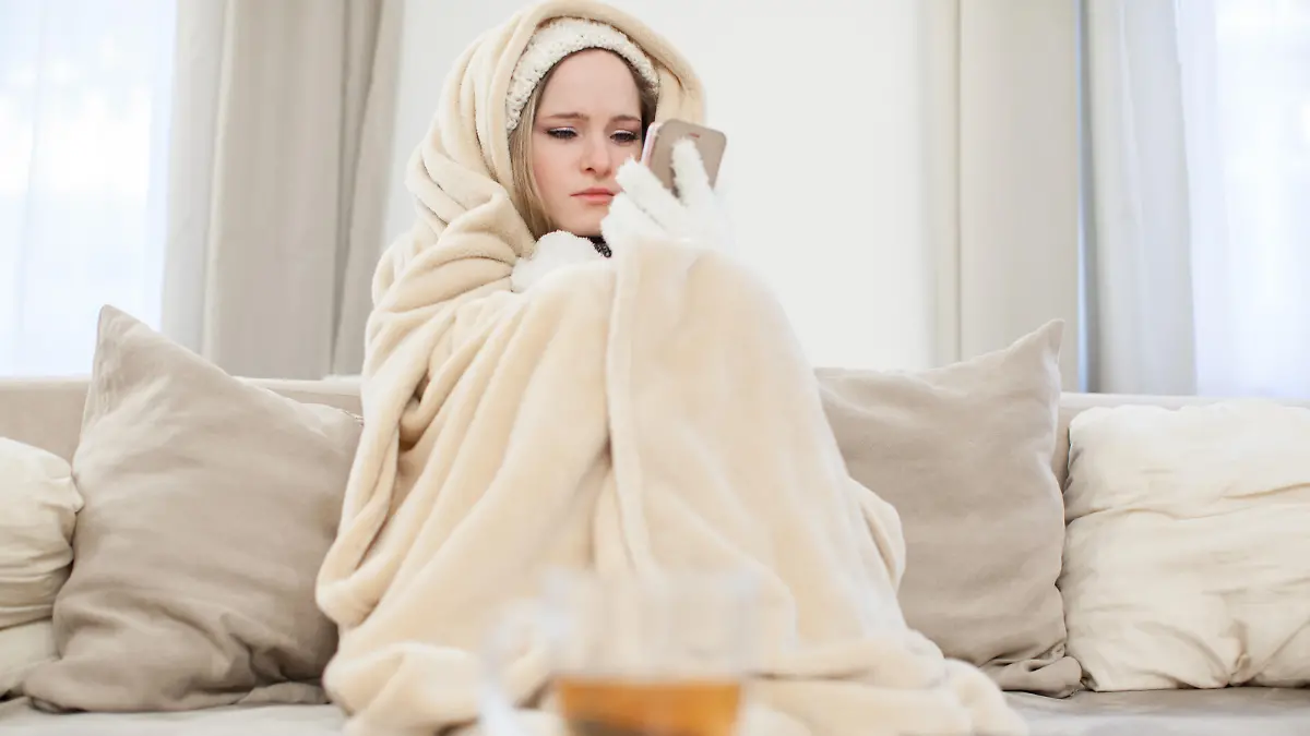 ILLUSTRATION - Eine junge Frau sitzt am 29.11.2016 in einem Wohnzimmer in Hamburg in Decken eingewickelt auf einer Couch und schaut auf ihr Handy, um den Vermieter zu informieren (gestellte Szene). Foto: Christin Klose || Modellfreigabe vorhanden