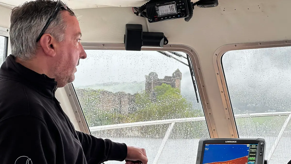 26.08.2023, Großbritannien, Drumnadrochit: Skipper Ali Matheson steht auf der Brücke eines Bootes auf dem Loch Ness, im Hintergrund ist die Ruine von Urquhart Castle zu sehen. In Schottland hat am Samstag die vermutlich größte Suche nach dem Ungeheuer von Loch Ness, genannt Nessie, seit Jahrzehnten begonnen. Dutzende Freiwillige aus aller Welt postierten sich am Samstagmorgen bei teils strömendem Regen an 17 Beobachtungsposten rund um den berühmten See in den Highlands. Tagsüber sollten Boote mit spezieller Technik wie einem Hydrophon - einer Art Unterwassermikrofon - über den Loch Ness kreuzen. Auch der Einsatz von Drohnen mit Wärmebildkameras war geplant. (zu dpa ««Größte Nessie-Suche seit Jahrzehnten» beginnt in Schottland») Foto: Benedikt von Imhoff/dpa +++ dpa-Bildfunk +++