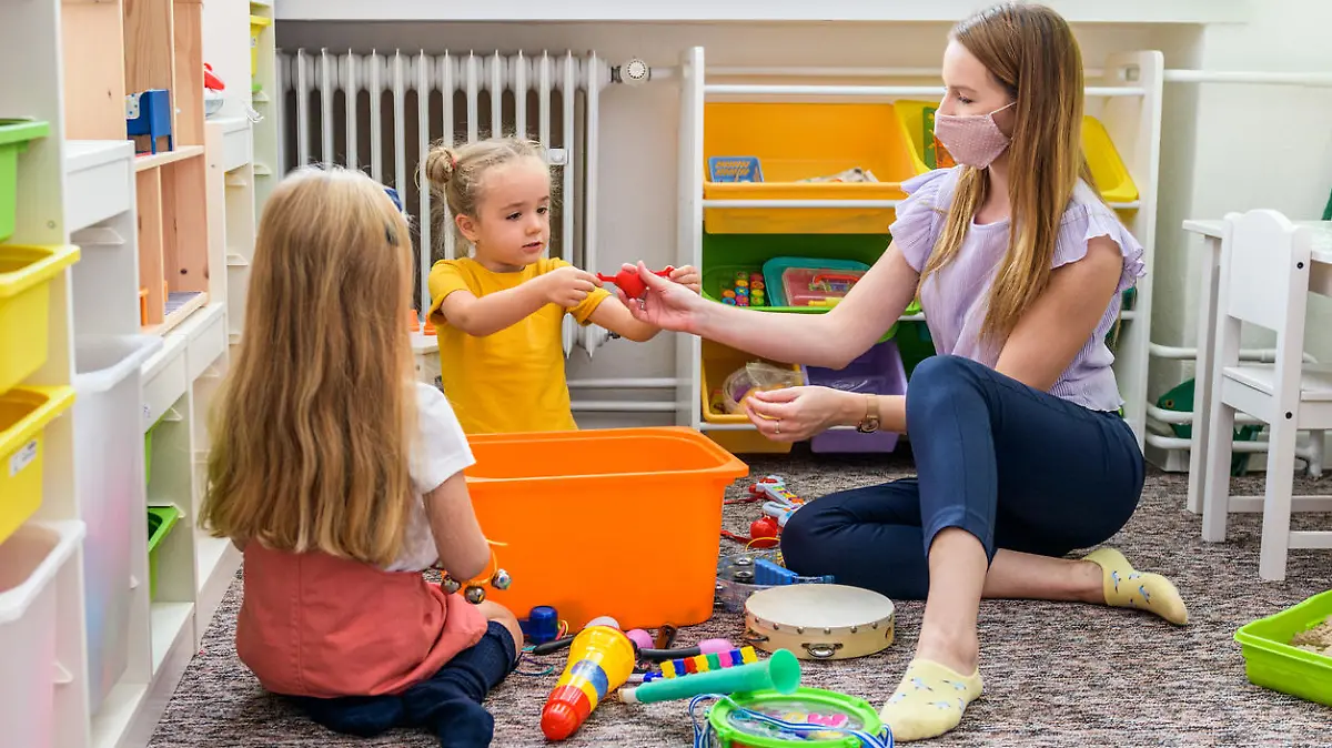 Young female therapist wearing protective face mask playing with two toddler girls during occupational child therapy.