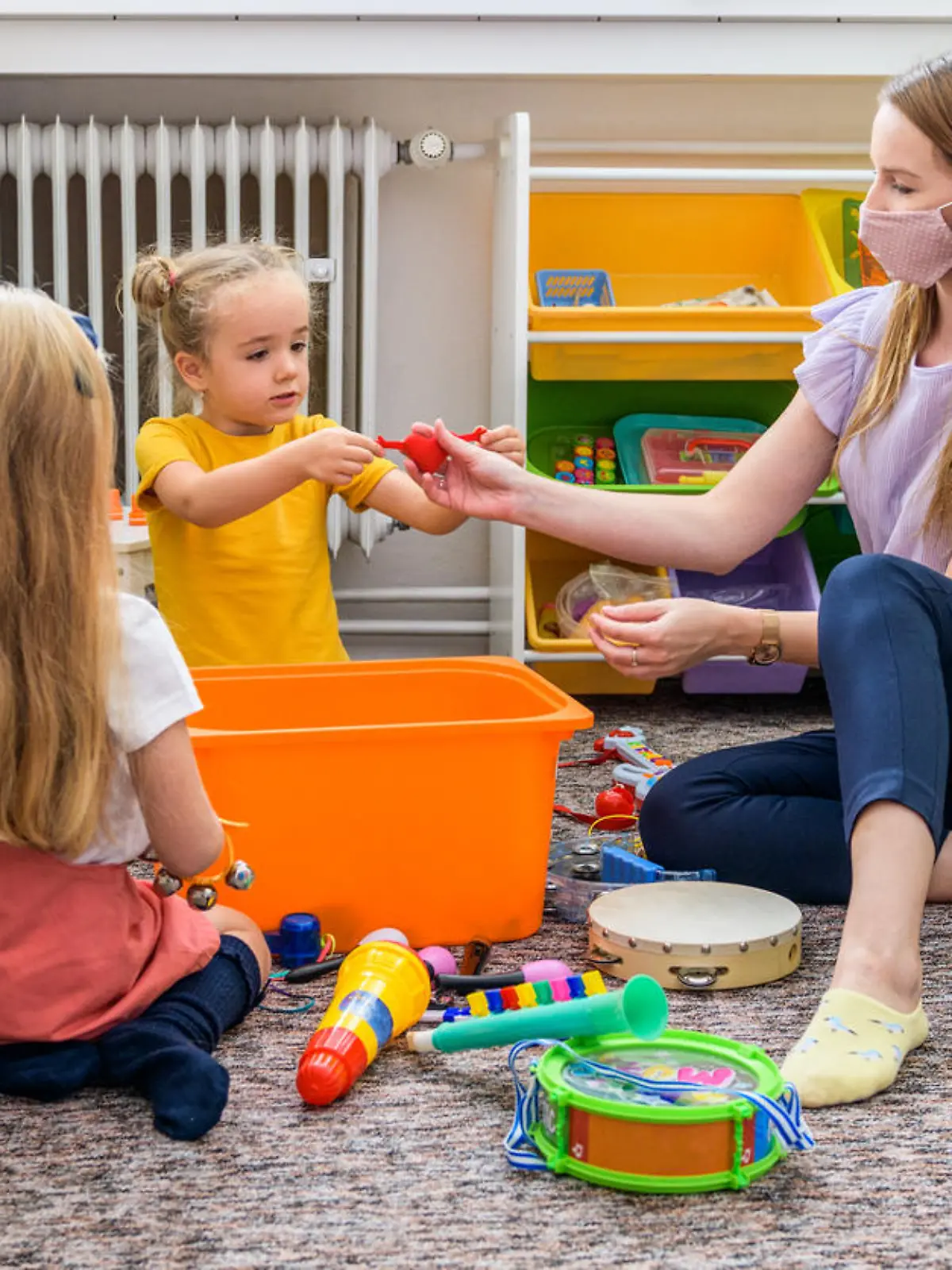 Young female therapist wearing protective face mask playing with two toddler girls during occupational child therapy.