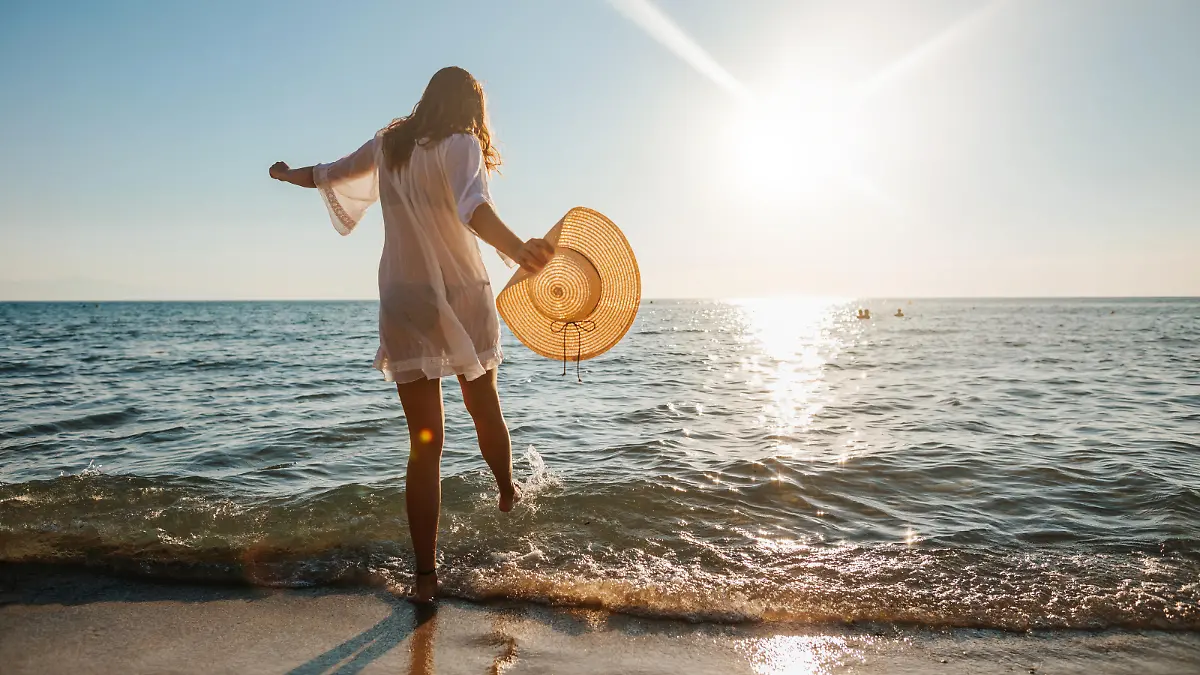 Close-up of young woman in white sun dress and with hat in hand walking alone on sandy beach at summer sunset, splashing water in sea shallow