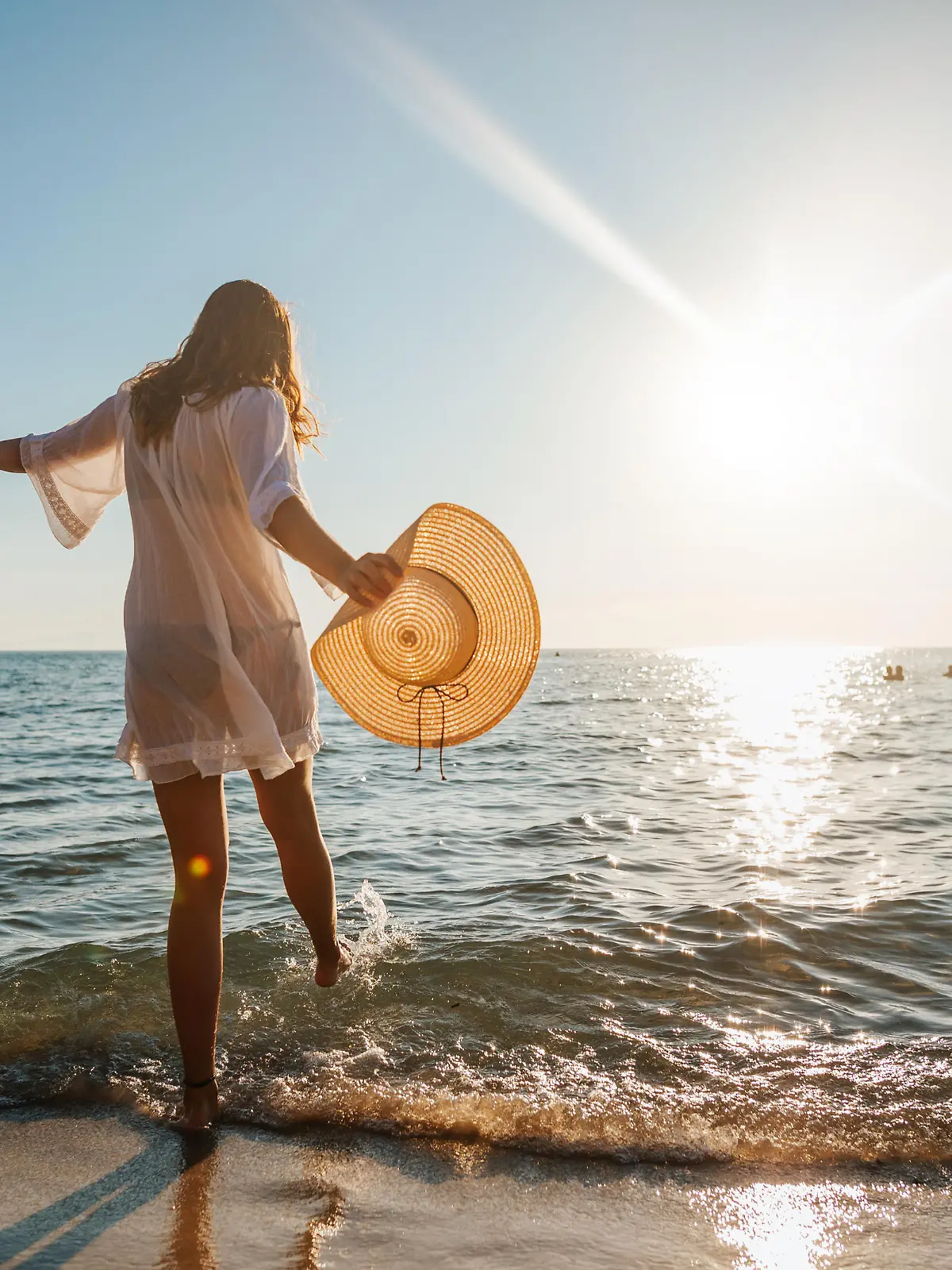 Close-up of young woman in white sun dress and with hat in hand walking alone on sandy beach at summer sunset, splashing water in sea shallow