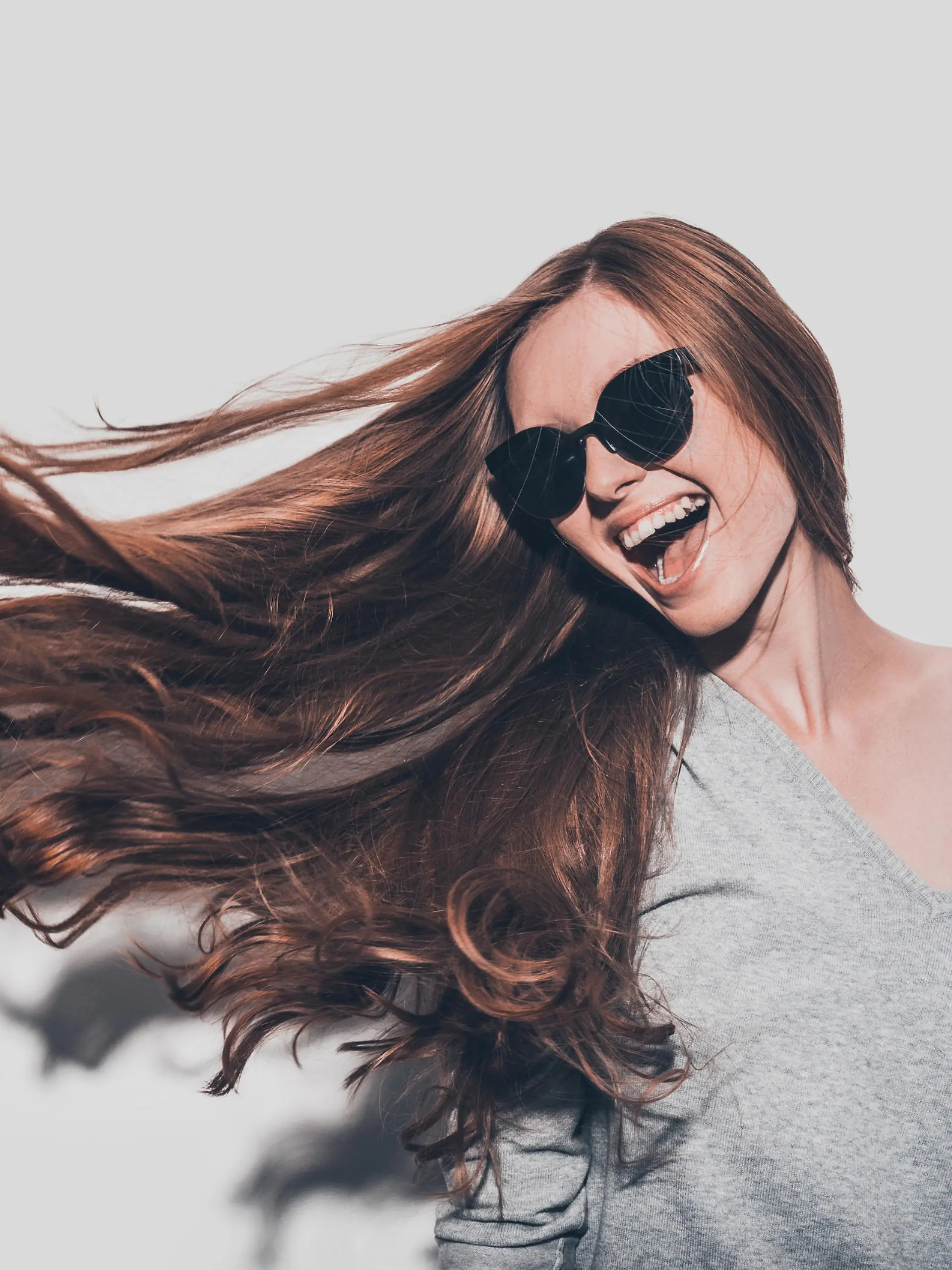 Attractive young smiling woman in sunglasses and with tousled hair looking away while standing against grey background
