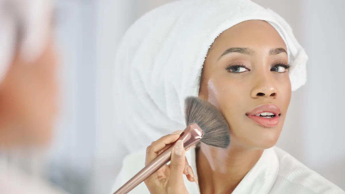 Attractive young Black woman in bathrobe and towel on her head applying make-up in front of mirror