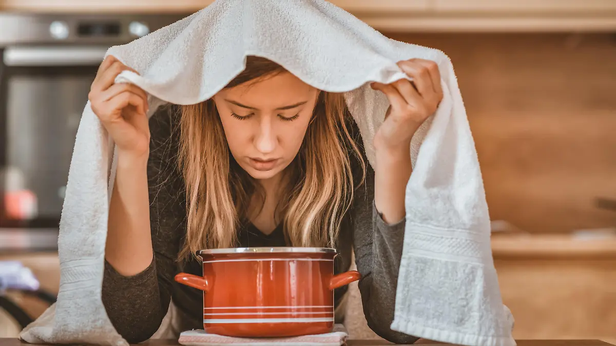 Health care, flu, hygiene and people concept - Photo of sick brown hair woman at home inhaling over a bowl. Rhinitis treatment at home by inhalation. Young woman inhaling chamomile tea steam as an alternative therapy or traditional cure. Young woman inhales steam to treat a blocked nose during the day.