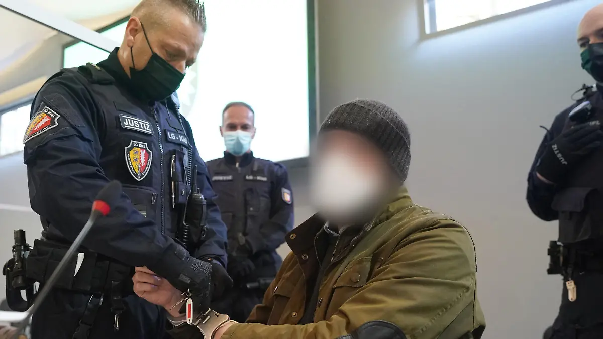 ARCHIV - 07.03.2022, Schleswig-Holstein, Kiel: Ein Justizbeamter entfernt die Handschellen des Angeklagten (2.v.r) im Sitzungssaal der Außenstelle des Landgerichts zu Beginn des Prozesstages wegen eines Dreifachmordes. Am 19. Mai 2021 soll der Zahnarzt zuerst seine von ihm getrennt lebende Ehefrau und deren neuen Bekannten in Dänischenhagen mit einer Maschinenpistole erschossen und danach in Kiel einen gemeinsamen Bekannten des Ehepaares mit einer halbautomatischen Pistole getötet haben. (zu dpa «Urteil erwartet in Dreifachmord-Prozess gegen Zahnarzt») Foto: Marcus Brandt/dpa - ACHTUNG: Der Angeklagte wurde aus rechtlichen Gründen gepixelt +++ dpa-Bildfunk +++