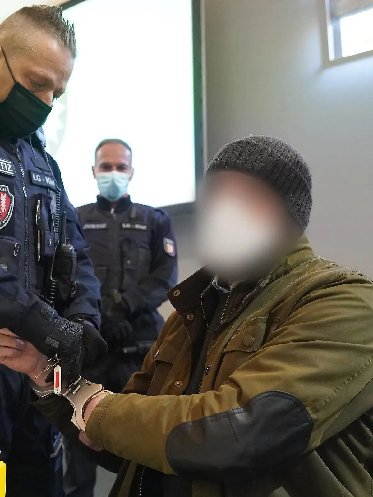 ARCHIV - 07.03.2022, Schleswig-Holstein, Kiel: Ein Justizbeamter entfernt die Handschellen des Angeklagten (2.v.r) im Sitzungssaal der Außenstelle des Landgerichts zu Beginn des Prozesstages wegen eines Dreifachmordes. Am 19. Mai 2021 soll der Zahnarzt zuerst seine von ihm getrennt lebende Ehefrau und deren neuen Bekannten in Dänischenhagen mit einer Maschinenpistole erschossen und danach in Kiel einen gemeinsamen Bekannten des Ehepaares mit einer halbautomatischen Pistole getötet haben. (zu dpa «Urteil erwartet in Dreifachmord-Prozess gegen Zahnarzt») Foto: Marcus Brandt/dpa - ACHTUNG: Der Angeklagte wurde aus rechtlichen Gründen gepixelt +++ dpa-Bildfunk +++