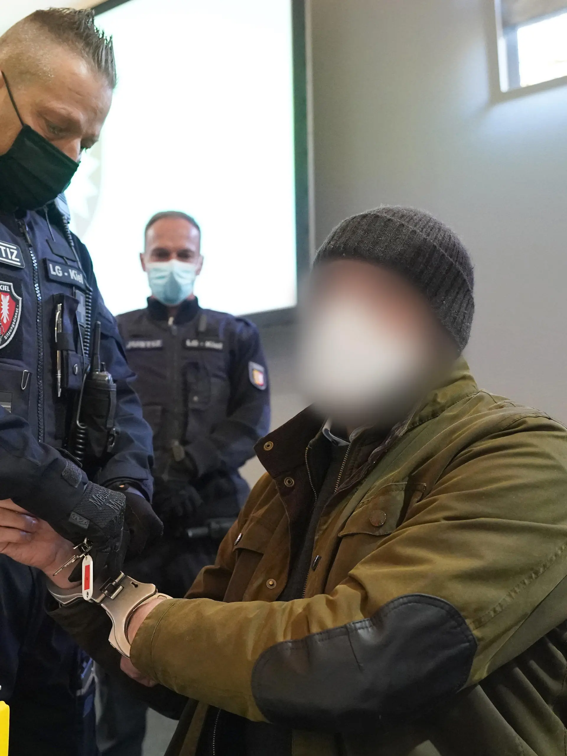 ARCHIV - 07.03.2022, Schleswig-Holstein, Kiel: Ein Justizbeamter entfernt die Handschellen des Angeklagten (2.v.r) im Sitzungssaal der Außenstelle des Landgerichts zu Beginn des Prozesstages wegen eines Dreifachmordes. Am 19. Mai 2021 soll der Zahnarzt zuerst seine von ihm getrennt lebende Ehefrau und deren neuen Bekannten in Dänischenhagen mit einer Maschinenpistole erschossen und danach in Kiel einen gemeinsamen Bekannten des Ehepaares mit einer halbautomatischen Pistole getötet haben. (zu dpa «Urteil erwartet in Dreifachmord-Prozess gegen Zahnarzt») Foto: Marcus Brandt/dpa - ACHTUNG: Der Angeklagte wurde aus rechtlichen Gründen gepixelt +++ dpa-Bildfunk +++