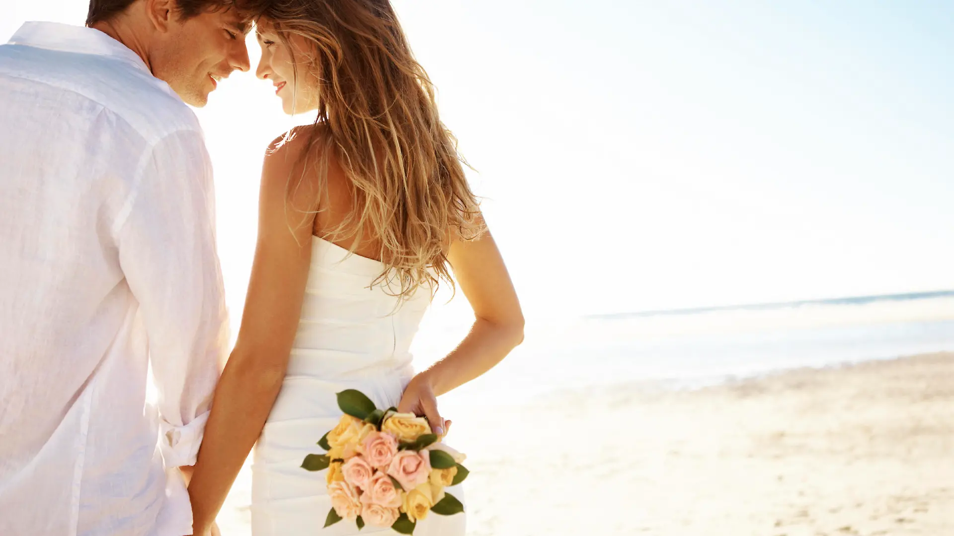 A young couple standing head-to-head on the beach while the woman holds flowers