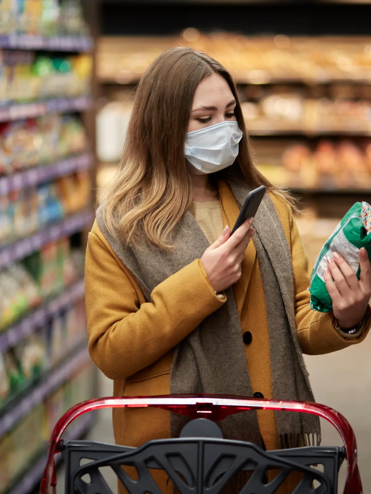 A young woman in a medical mask with a smartphone in her hand scans the product. There is a large shopping cart nearby.