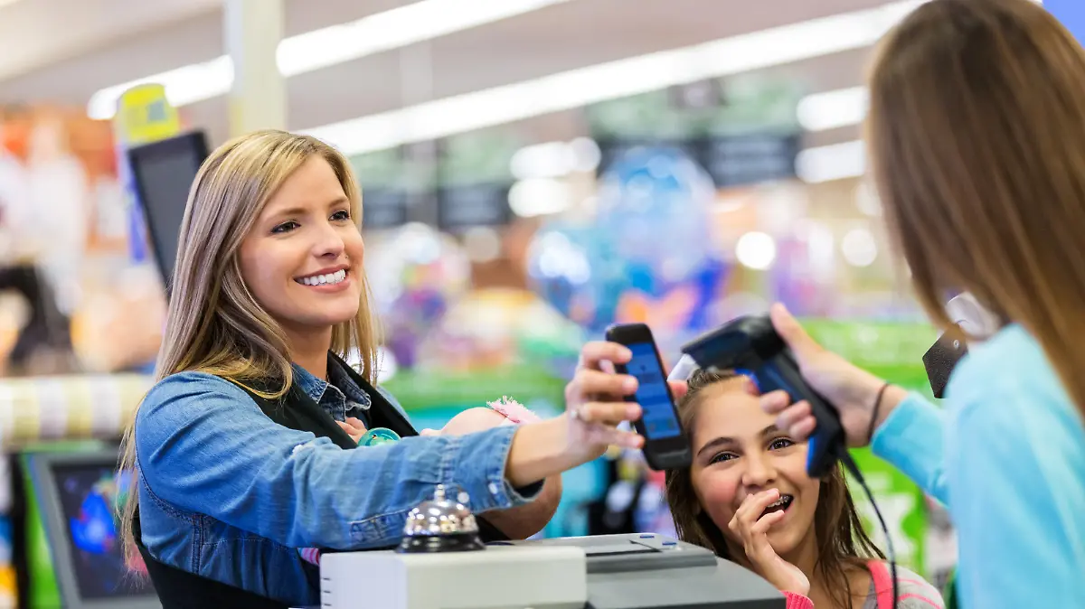 Mid adult Caucasian woman is using her smart phone app to pay for groceries in local grocery store. Employee is scanning smart phone with scanner. Woman is shopping with elementary age daughter and four month old baby. She is wearing her baby in a mei tai style baby wearing wrap.