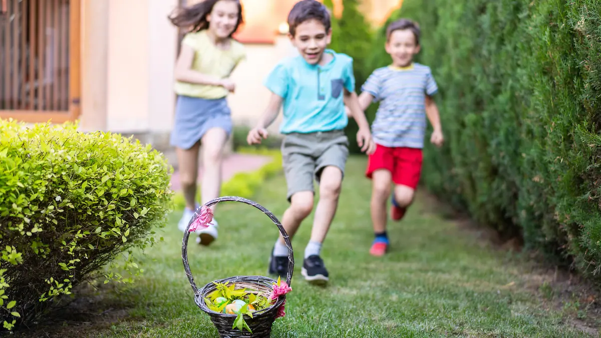 Excited children are running towards a decorated basket filled with Easter eggs sitting on the ground in a garden on a bright sunny day