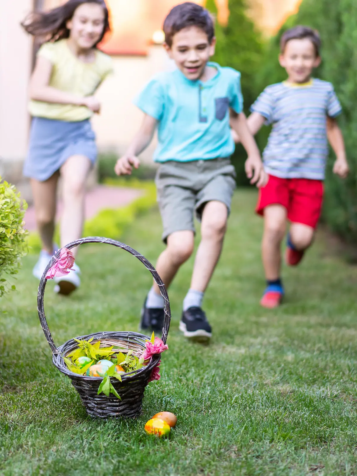 Excited children are running towards a decorated basket filled with Easter eggs sitting on the ground in a garden on a bright sunny day