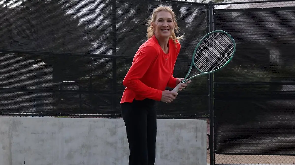 Steffi Graf steht mit Tennisschläger auf einem Tennisplatz und lacht seitlich in die Kamera
