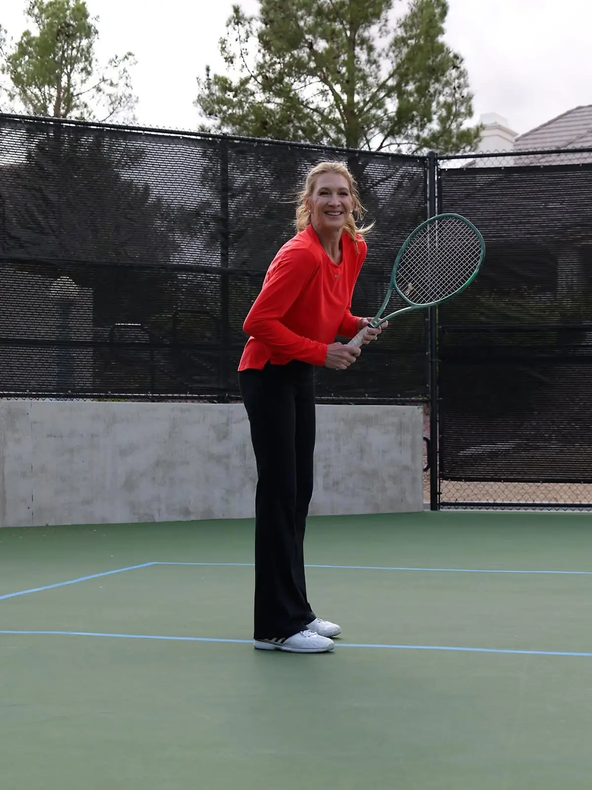 Steffi Graf steht mit Tennisschläger auf einem Tennisplatz und lacht seitlich in die Kamera