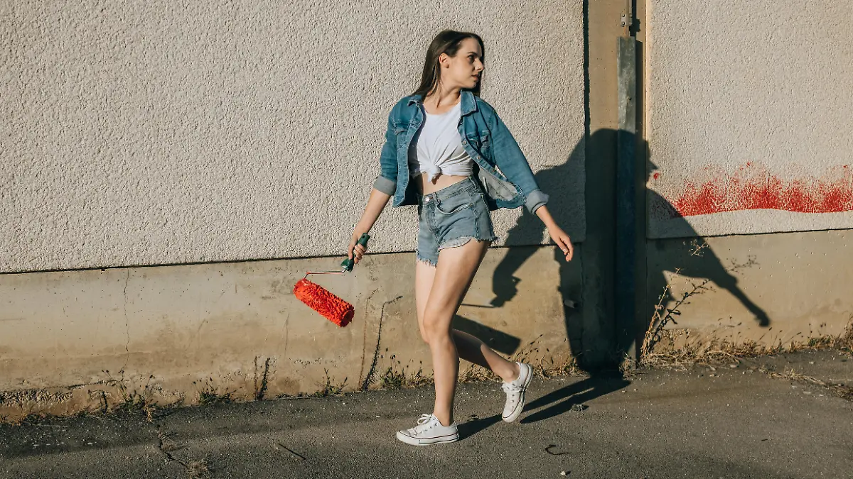 Beautiful young woman holding red paint roller while walking beside concrete wall
