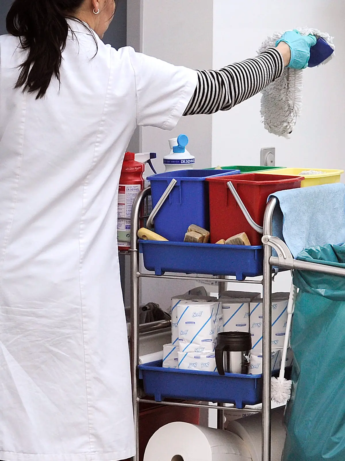 Eine Putzkraft steht arbeitet an einem Reinigungswagen. Die Frau hat einen Lappen in der Hand.