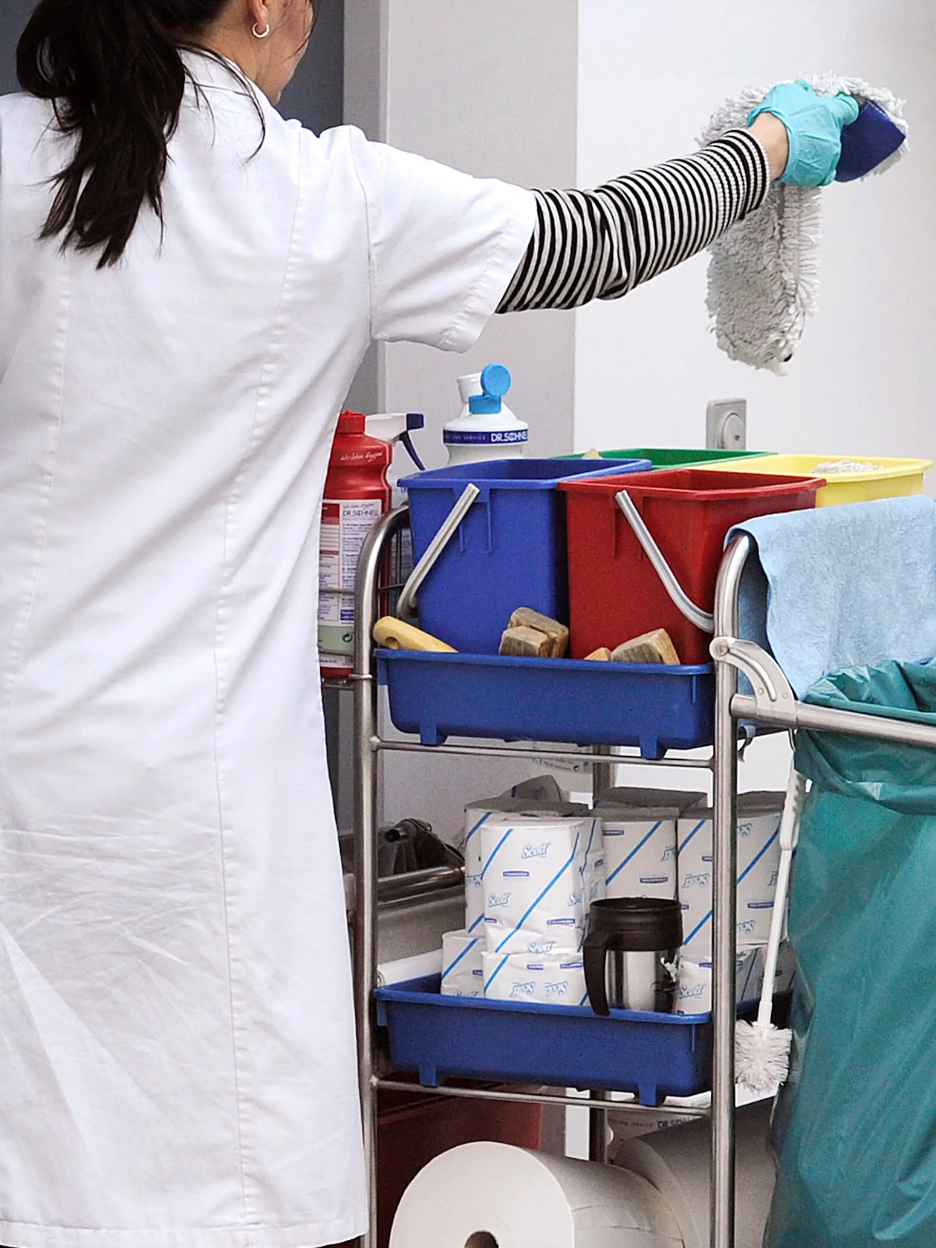 Eine Putzkraft steht arbeitet an einem Reinigungswagen. Die Frau hat einen Lappen in der Hand.