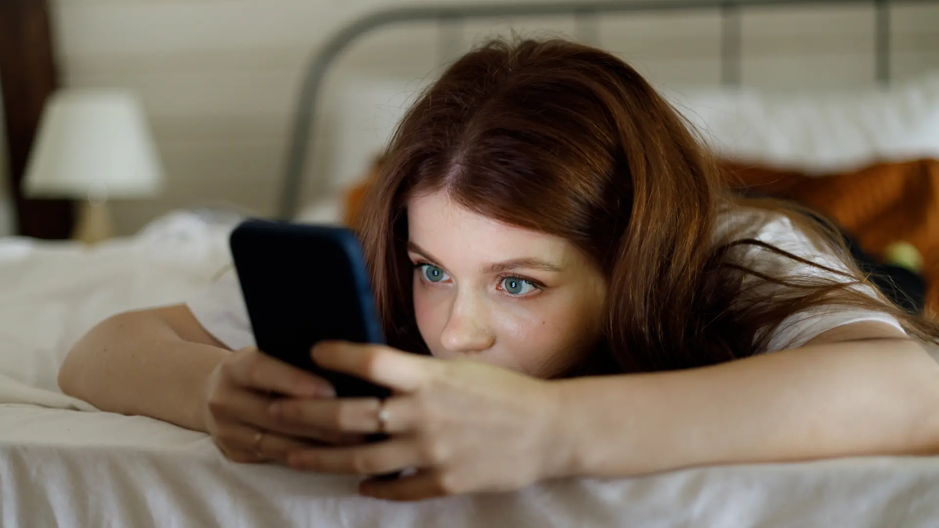 Eine junge Frau liegt auf dem Bett und surft am Handy.