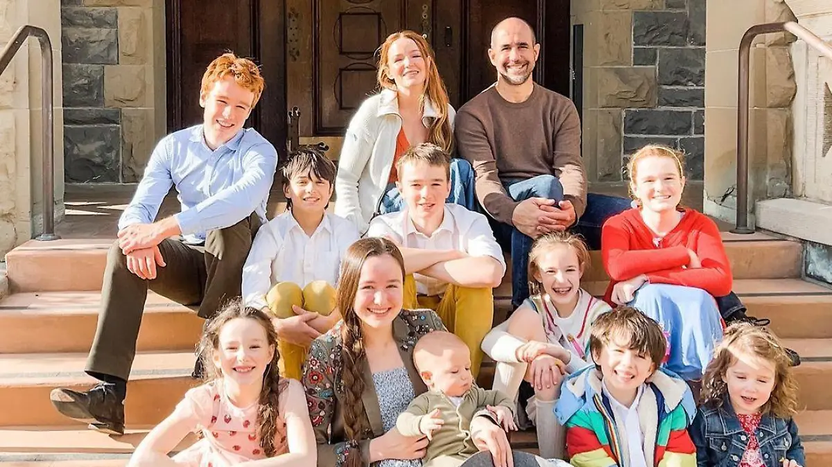 Amber de la Motte mit ihrem Mann und ihren zwölf gemeinsamen Kindern sitzen auf einer Treppe vor einem Gebäude.