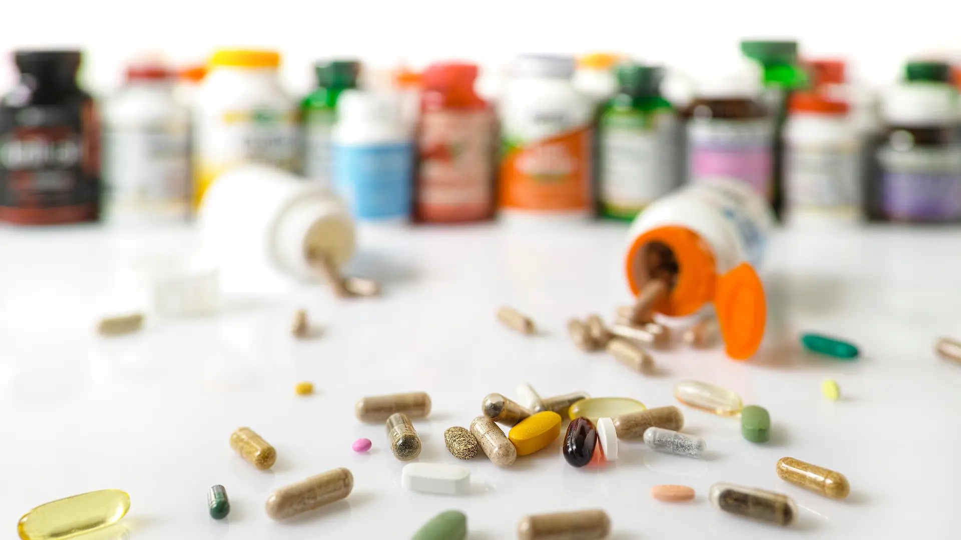 Colorful display and stack of many types of nutritional supplements and vitamin plastic bottles in an out of focus background with many supplement and vitamin capsules and pills in focus in the foreground. Scene set on a white background.