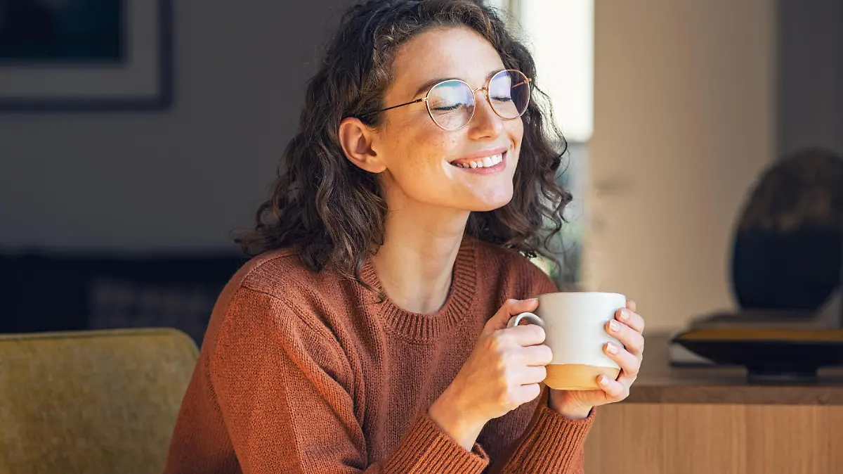 Happy young woman drinking a cup of tea in an autumn morning. Dreaming girl sitting in living room with cup of hot coffee enjoying under blanket with closed eyes. Pretty woman wearing sweater at home and enjoy a ray of sunshine on a winter afternoon.