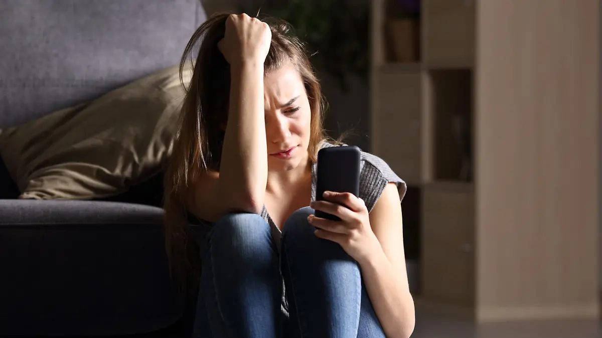Front view of a sad teen checking phone sitting on the floor in the living room at home with a dark background