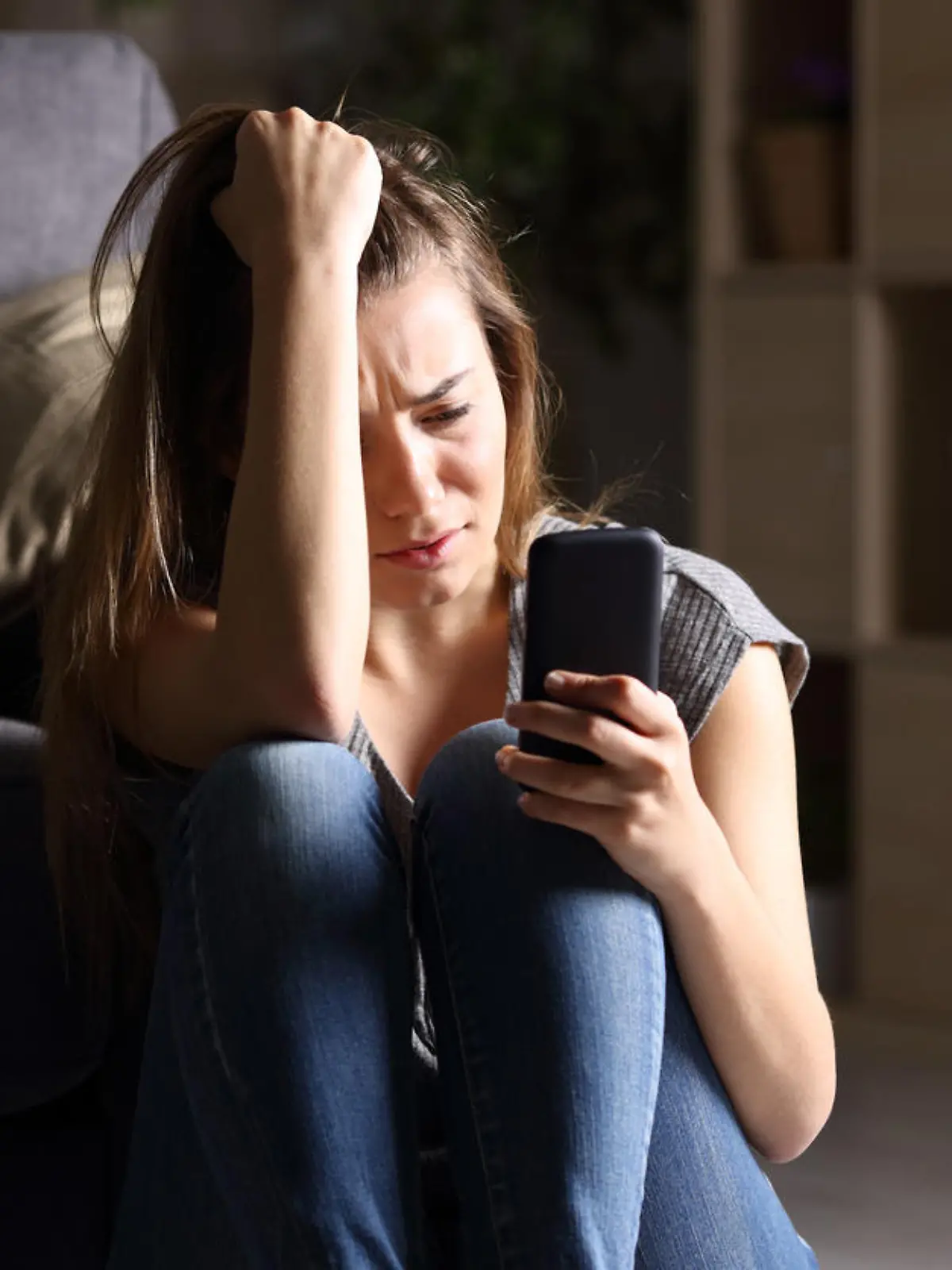 Front view of a sad teen checking phone sitting on the floor in the living room at home with a dark background