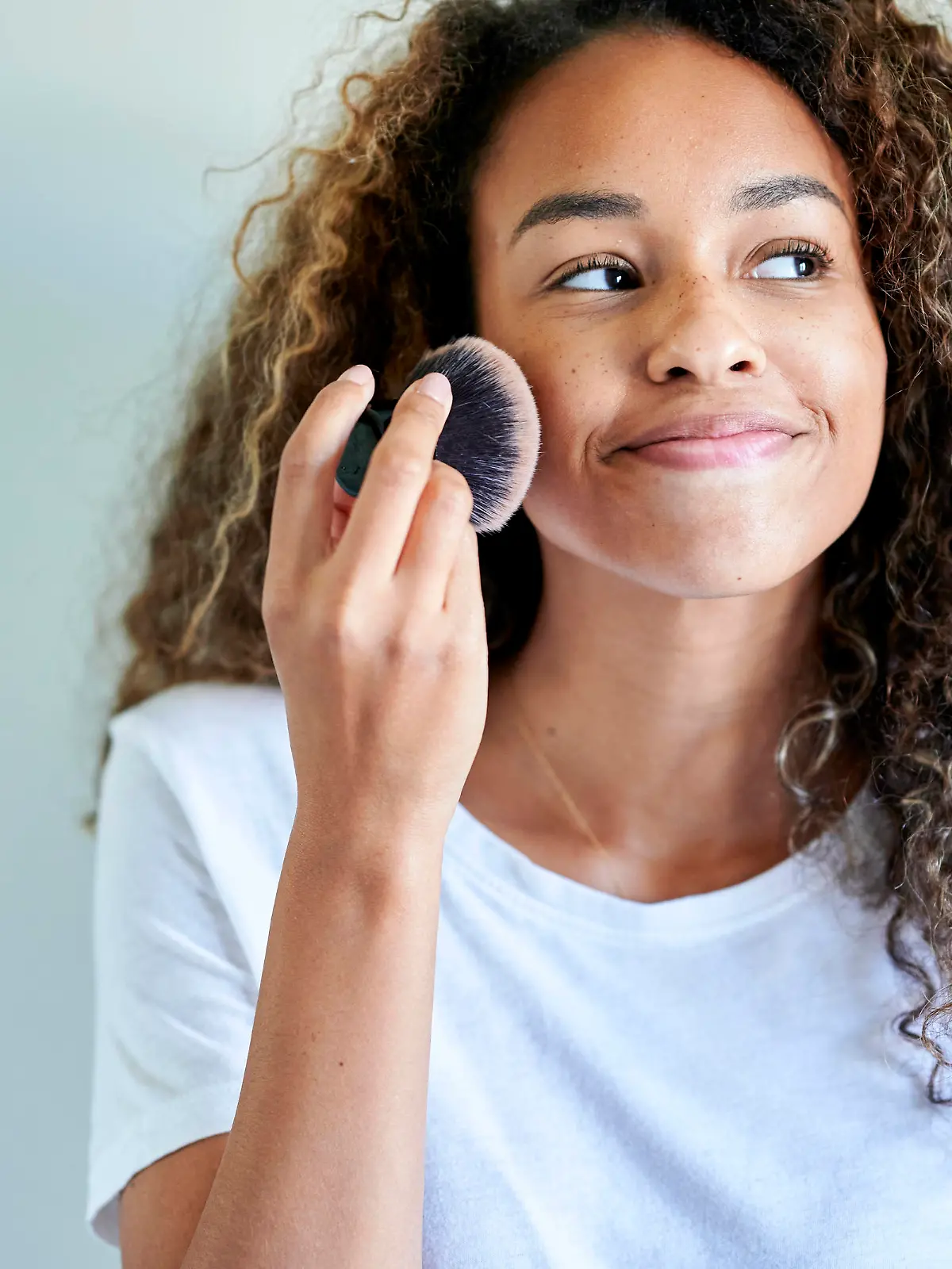 Smiling young woman applying face powder with make-up brush against white wall model released Symbolfoto KIJF03507 