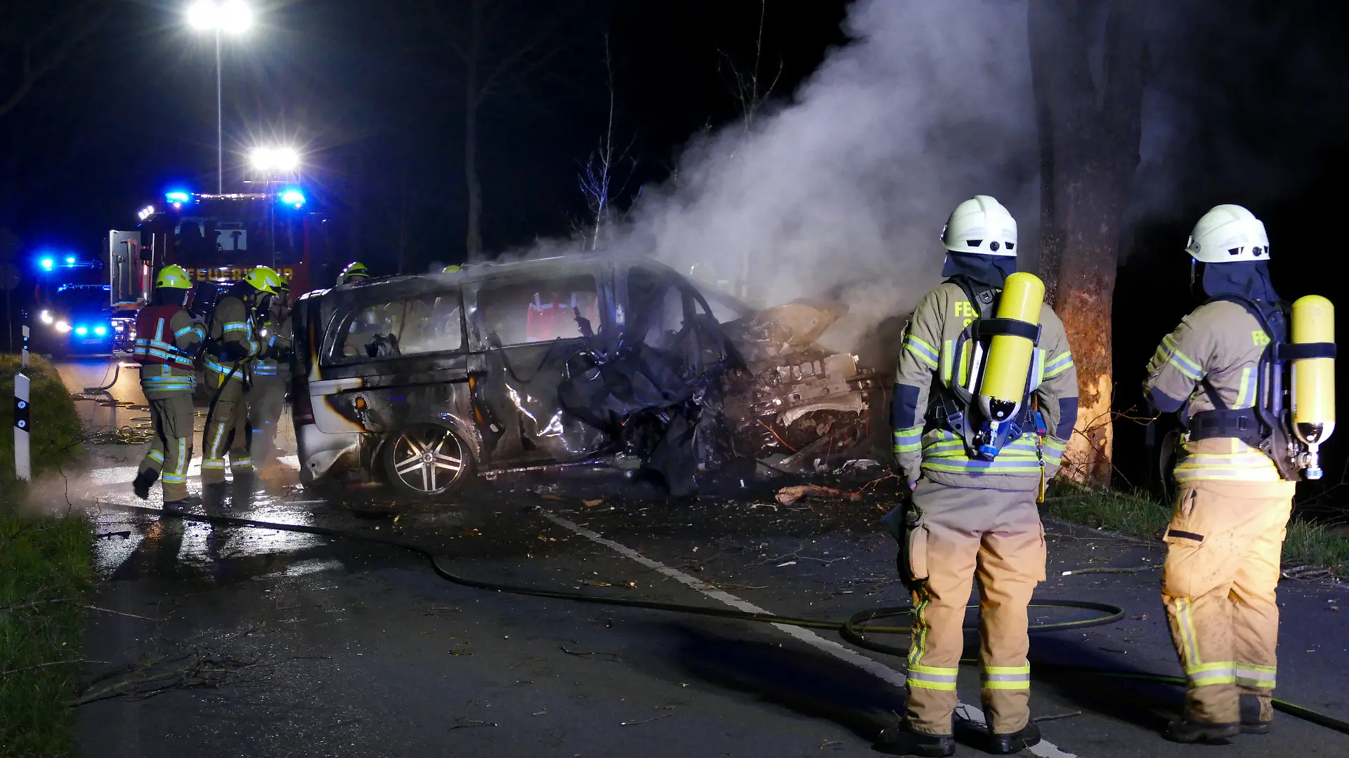 26.03.2022, Siegburg: Die Feuerwehr steht vor dem Wrack eines ausgebrannten Autos, in dem zwei Menschen ums Leben gekommen sind. Laut Polizei waren drei junge Männer in einem Lieferwagen in einer Kurve von der Straße abgekommen und gegen einen Baum geprallt. Der Wagen sei zurückgeschleudert und in Brand geraten, sagte der Sprecher. Die Getöteten seien junge Männer aus Siegburg des Jahrgangs 2005, sagte der Sprecher. Foto: Christof Schmoll/dpa +++ dpa-Bildfunk +++