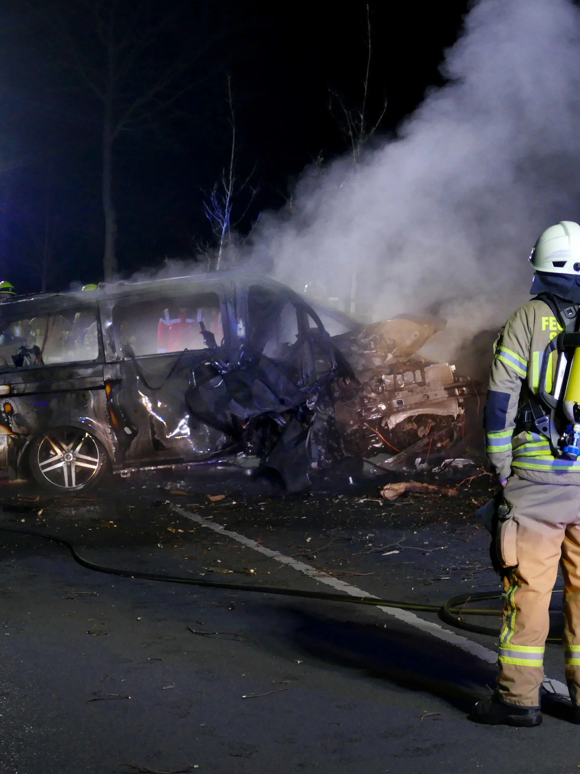 26.03.2022, Siegburg: Die Feuerwehr steht vor dem Wrack eines ausgebrannten Autos, in dem zwei Menschen ums Leben gekommen sind. Laut Polizei waren drei junge Männer in einem Lieferwagen in einer Kurve von der Straße abgekommen und gegen einen Baum geprallt. Der Wagen sei zurückgeschleudert und in Brand geraten, sagte der Sprecher. Die Getöteten seien junge Männer aus Siegburg des Jahrgangs 2005, sagte der Sprecher. Foto: Christof Schmoll/dpa +++ dpa-Bildfunk +++