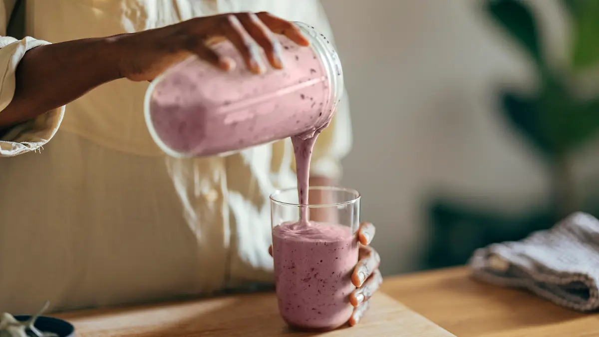 Close-up of hands of an Afro-American woman wearing beige shirt and pouring smoothie into a glass on a kitchen table. There is a part of a blender, a cutting board and a cloth on the desk in front of her. In the corner of a room sits a big plant.