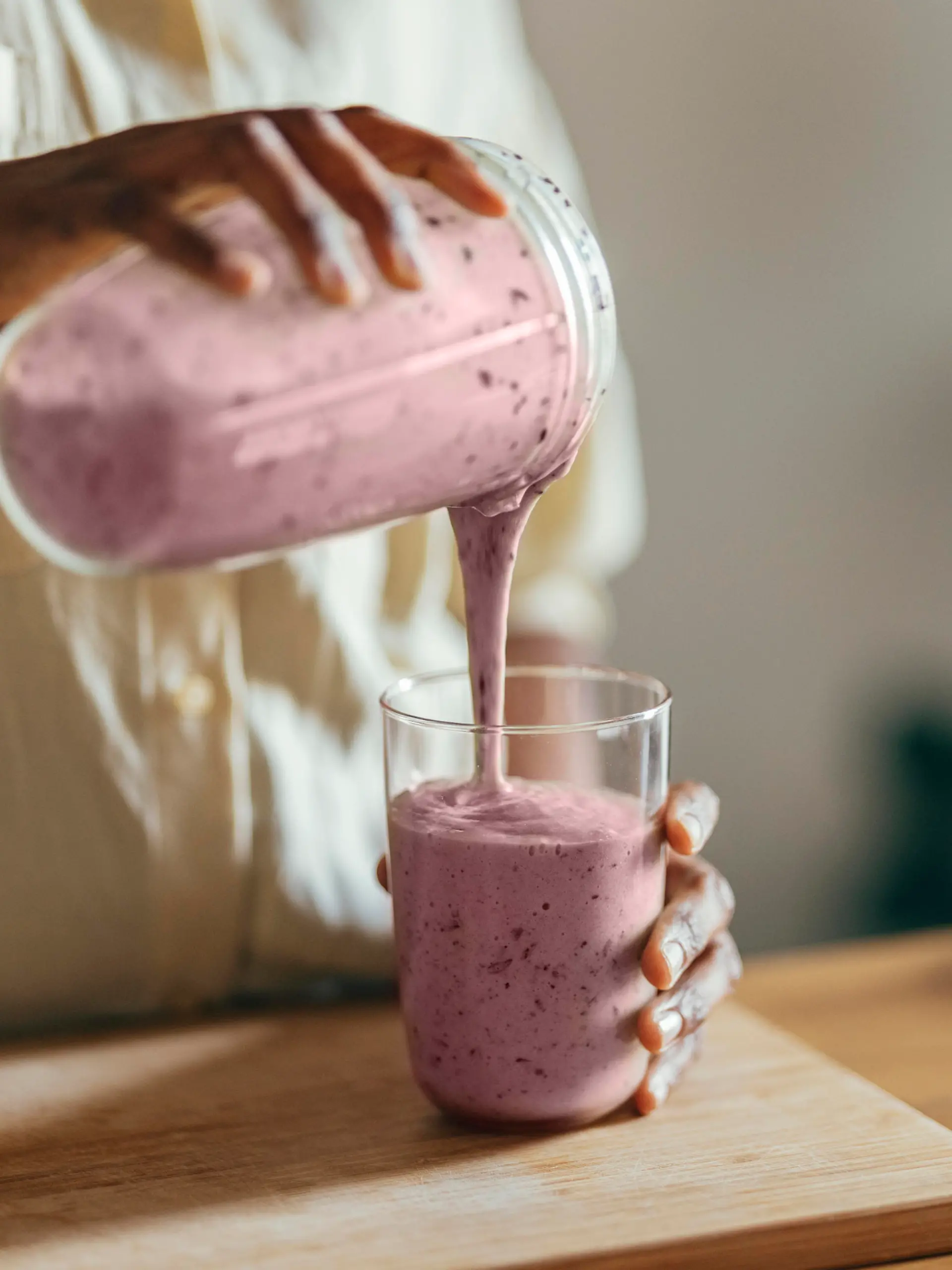 Close-up of hands of an Afro-American woman wearing beige shirt and pouring smoothie into a glass on a kitchen table. There is a part of a blender, a cutting board and a cloth on the desk in front of her. In the corner of a room sits a big plant.