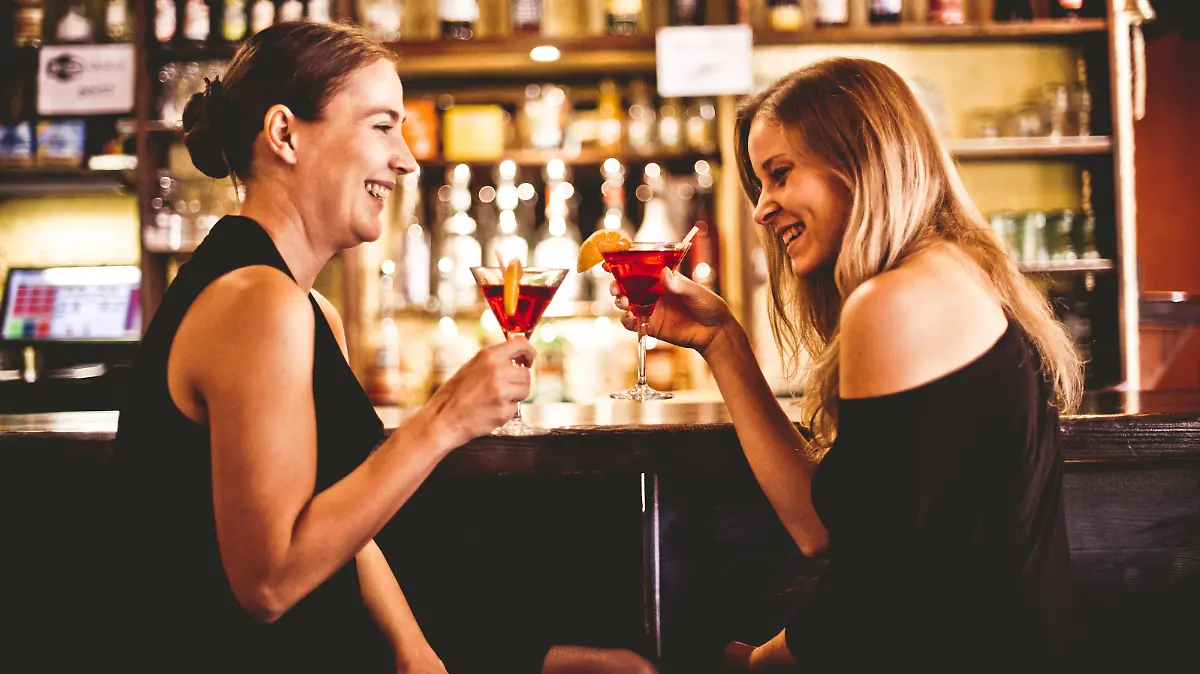 Two young smiling women sitting, drinking cocktail and having fun