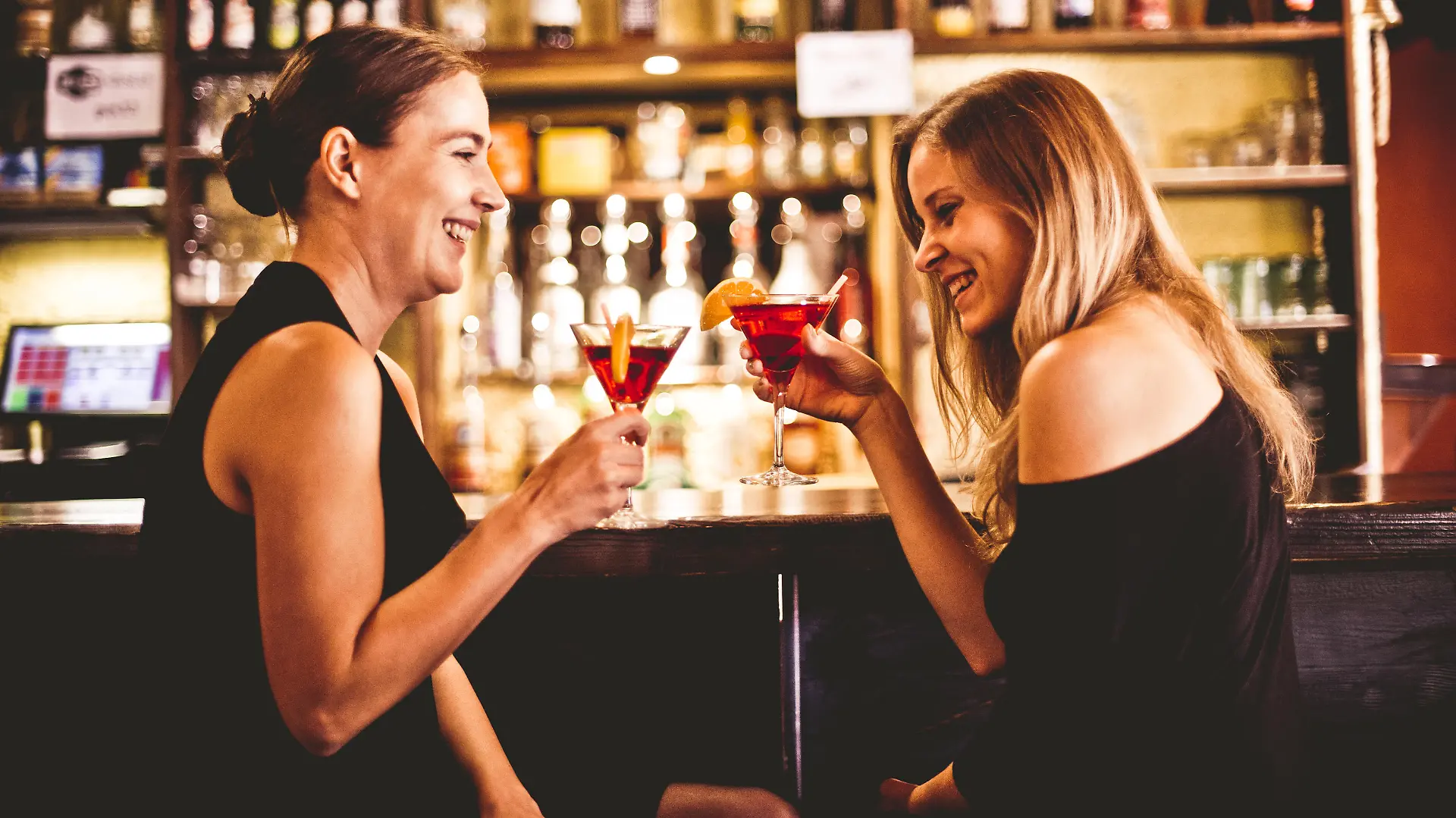 Two young smiling women sitting, drinking cocktail and having fun