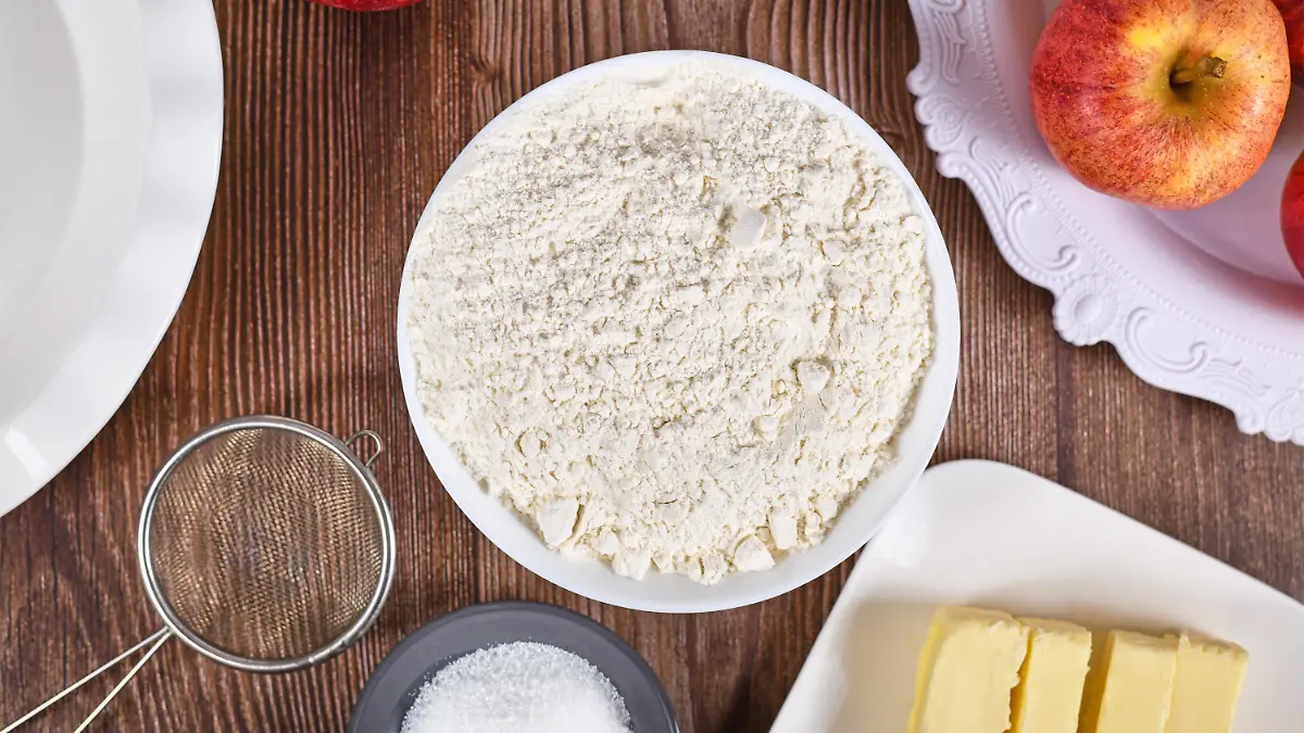 Bowl of flour surrounded by other pie crust ingredients like butter and sugar on wooden background