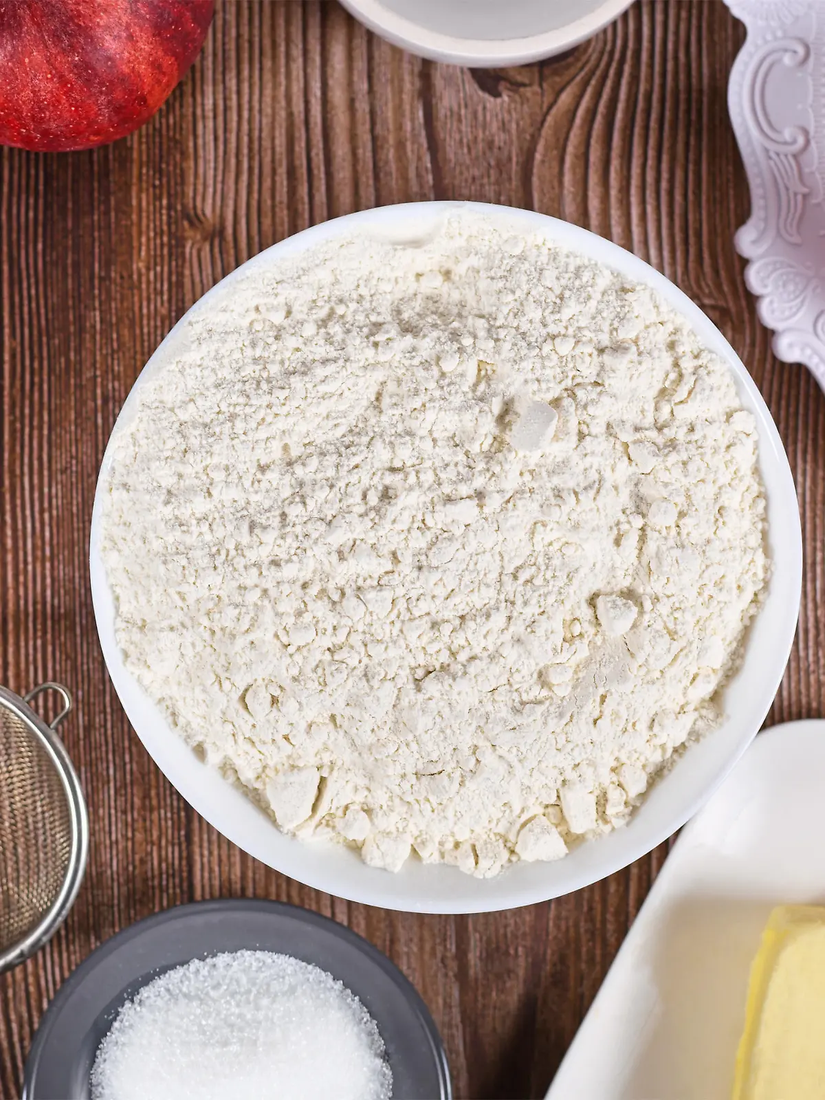 Bowl of flour surrounded by other pie crust ingredients like butter and sugar on wooden background
