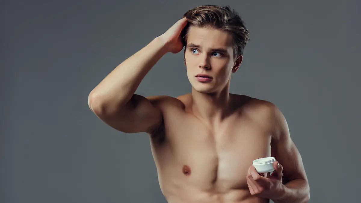 Handsome young man isolated. Portrait of shirtless muscular man is standing on grey background and using hair wax.