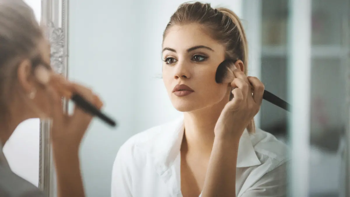 Closeup shot of attractive mid 20's blond woman putting on some makeup in the morning before going out. She's standing in front of big bathroom mirror, rear view.