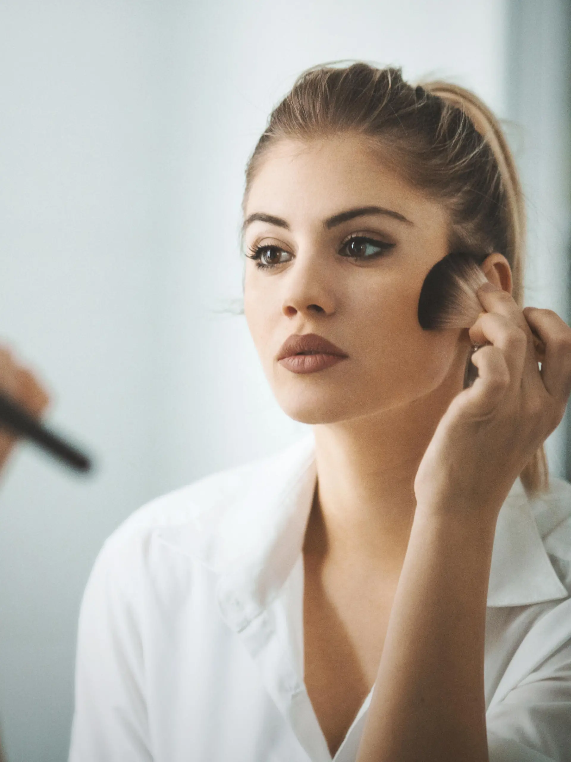 Closeup shot of attractive mid 20's blond woman putting on some makeup in the morning before going out. She's standing in front of big bathroom mirror, rear view.