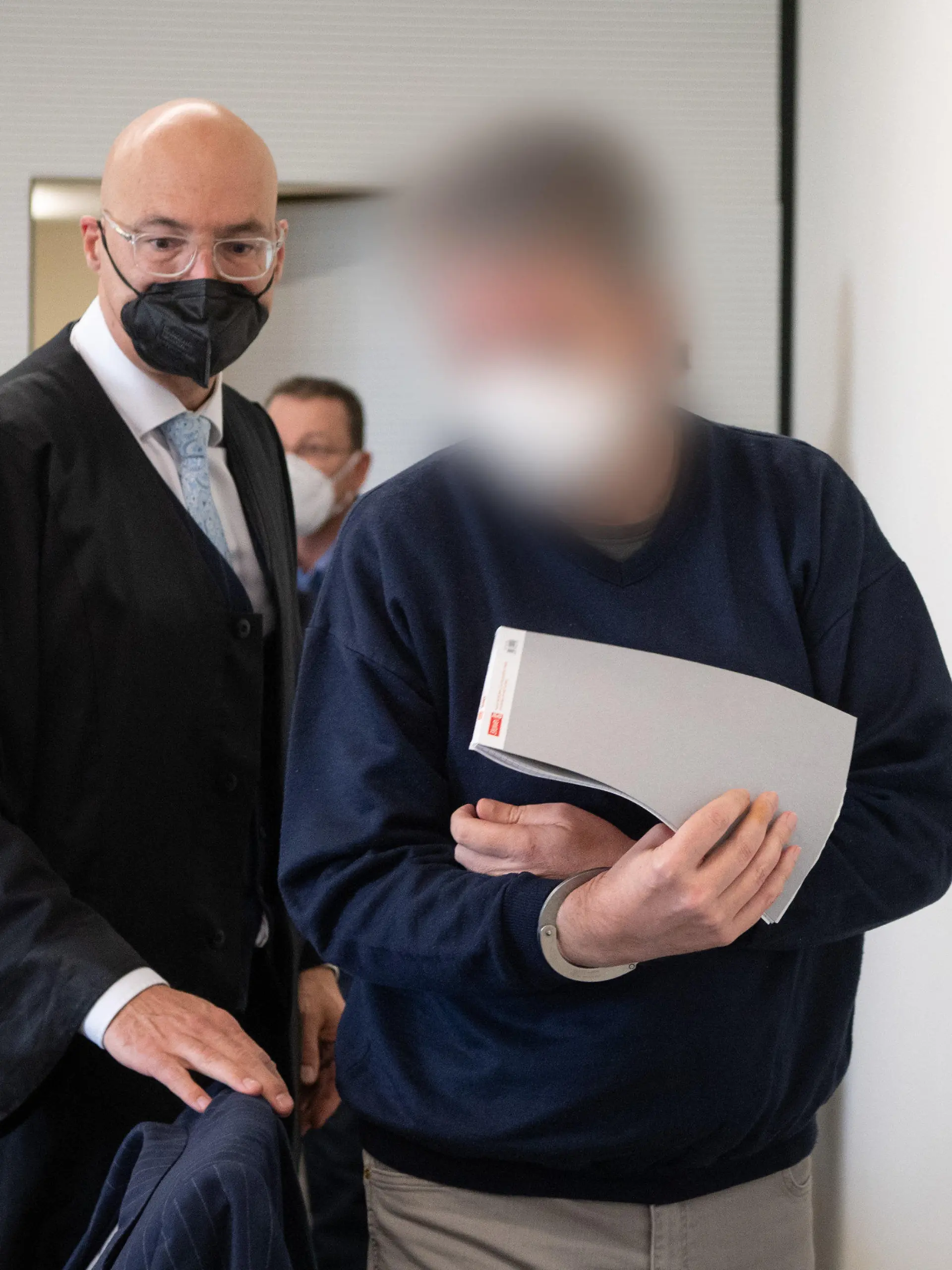 21.03.2022, Rheinland-Pfalz, Bad Kreuznach: Der Angeklagte wird in Handschellen in den Gerichtssaal gebracht. Links sein Anwalt Alexander Klein. Der Mann soll im vergangenen Jahr in Bad Kreuznach den Mitarbeiter einer Tankstelle erschossen haben, nachdem dieser ihn auf die coronabedingte Maskenpflicht hingewiesen hatte. Foto: Boris Roessler/dpa - ACHTUNG: Der Angeklagte wurde auf Anordnung des Gerichts gepixelt +++ dpa-Bildfunk +++