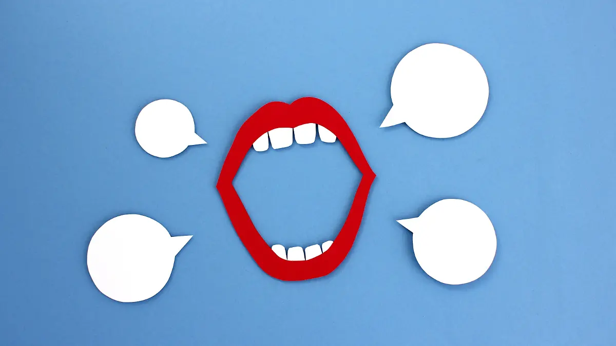 Paper cutout of open mouth with red lips and white teeth placed near empty speech balloons on blue background