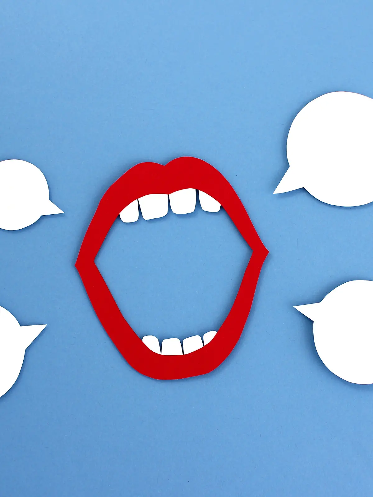 Paper cutout of open mouth with red lips and white teeth placed near empty speech balloons on blue background