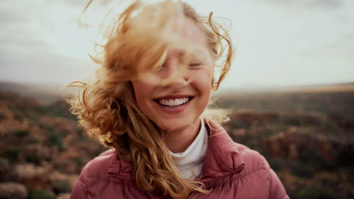 Portrait of young smiling woman face partially covered with flying hair standing at mountain - carefree woman